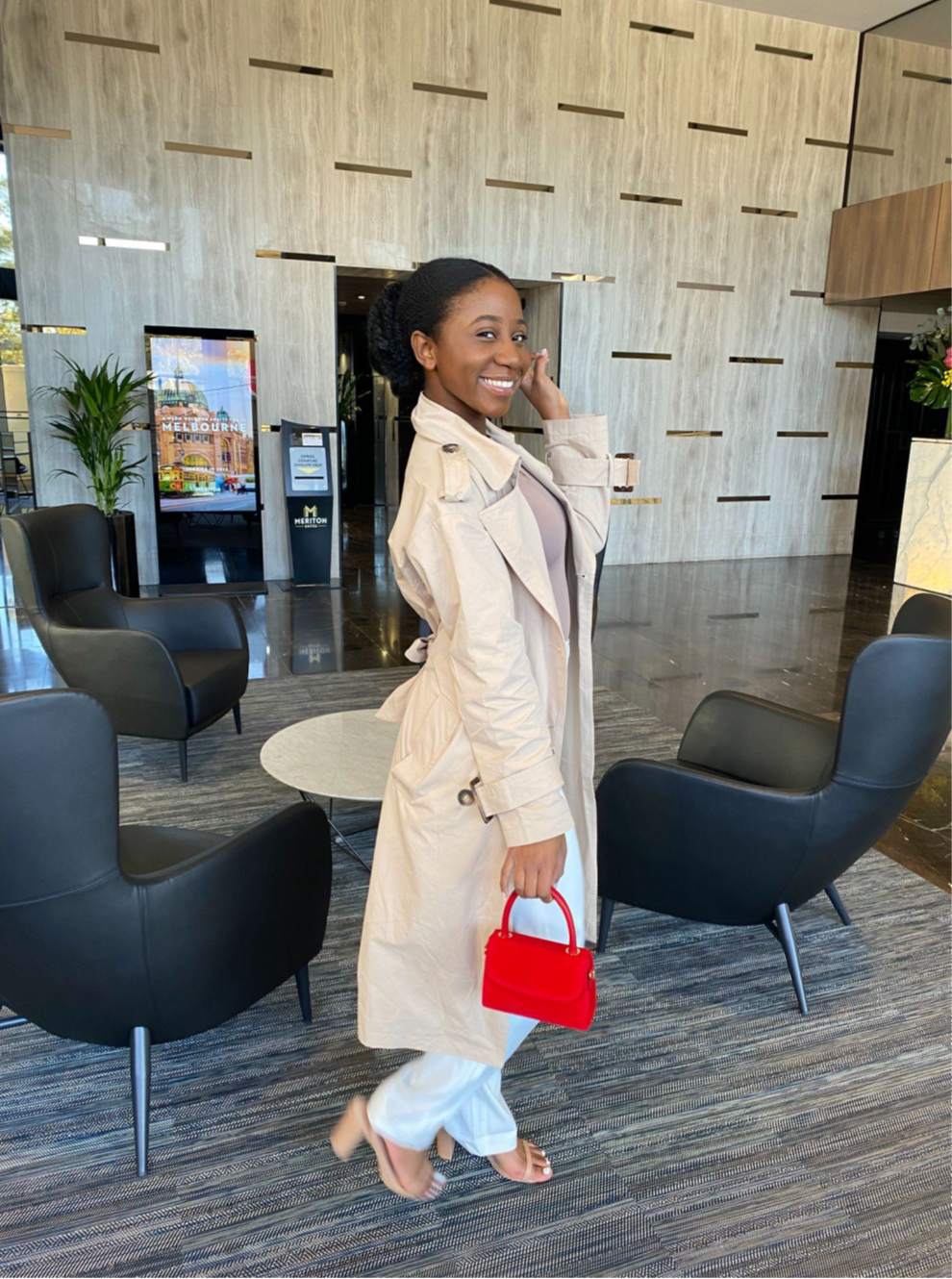 Photo of young woman smiling and waving, walking through a lounge area carrying red hand bag